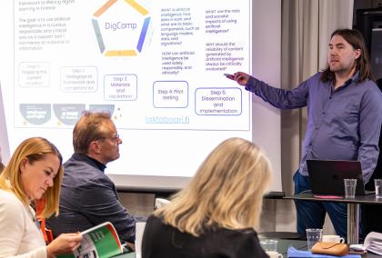 A man giving a presentation about Assistive Intelligence, with slides behind him and group of people listening to him.