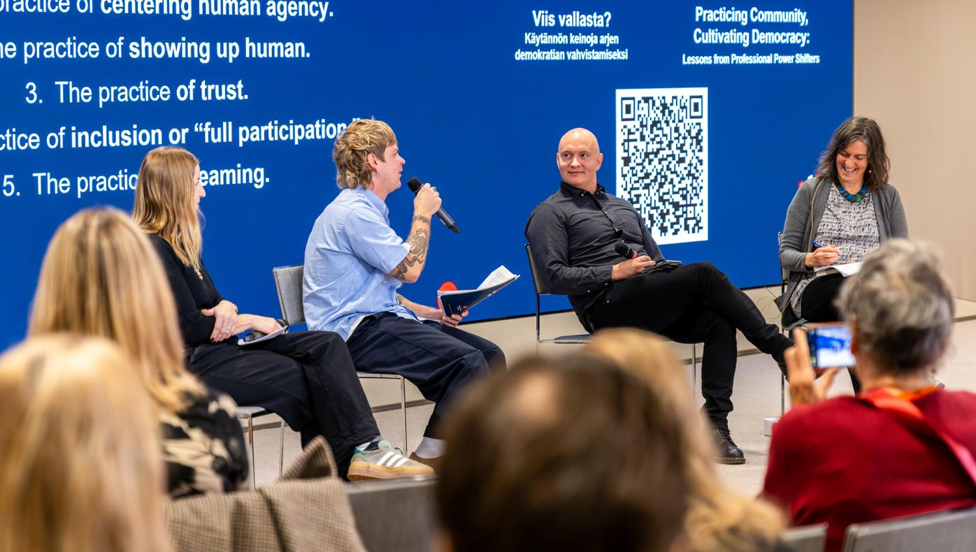Four panelists sitting in a row in front of a big screen. Person second from the left has the microphone and is enthusiastically speaking.