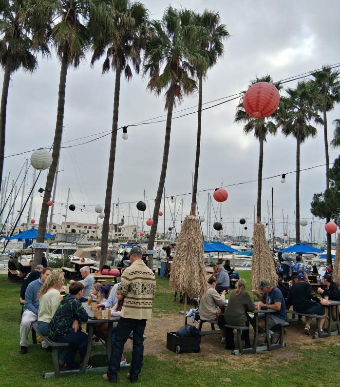 Photograph of an outdoor gathering at a marina with people seated at picnic tables under tall palm trees decorated with hanging lanterns in red, white, and black. Boats and sailboats are docked in the background
