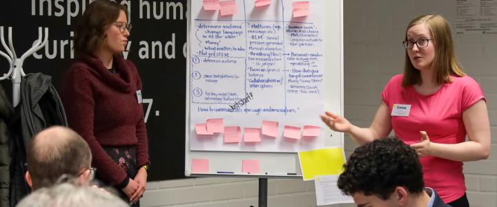 A person explaining what is written on a whiteboard next to them. The whiteboard includes colorful post-it notes and text.