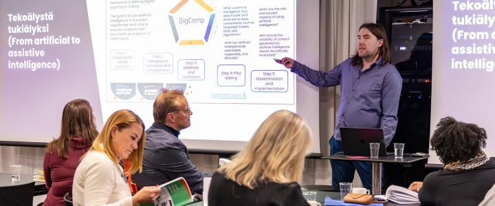 A man giving a presentation about Assistive Intelligence, with slides behind him and group of people listening to him.
