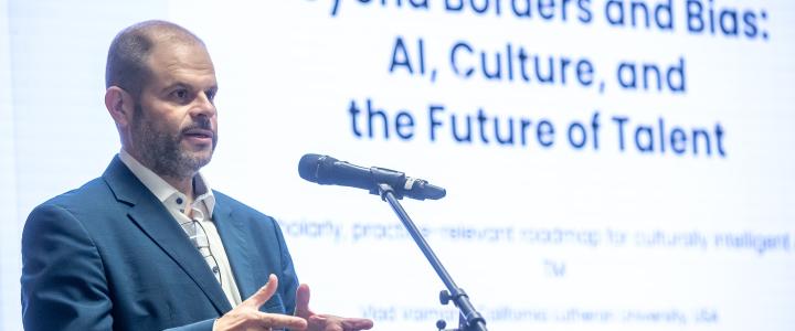 Vlad Vaiman giving a presentation, with a title slide behind him saying "Beyond borders and bias: AI, Culture, and the Future of Talent"